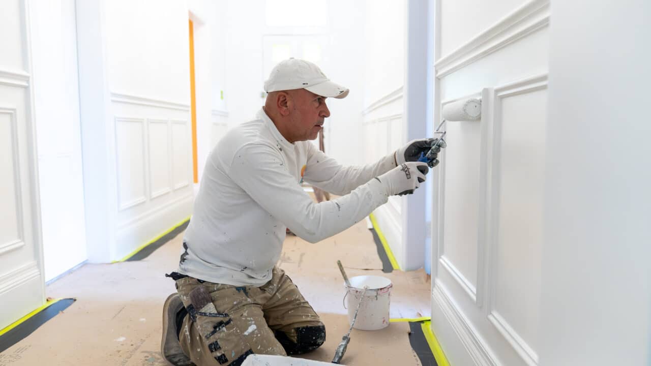 A person wearing a cap and overalls paints white paneling on a wall using a roller. The floor is covered for protection.