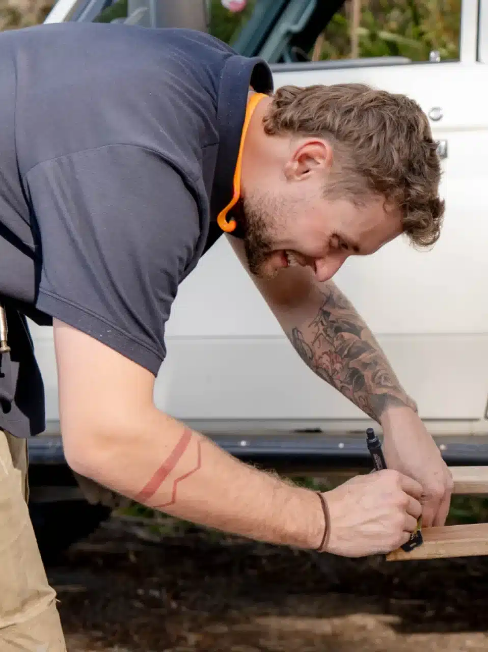A man with tattoos marks a piece of wood while outdoors, next to a white vehicle.