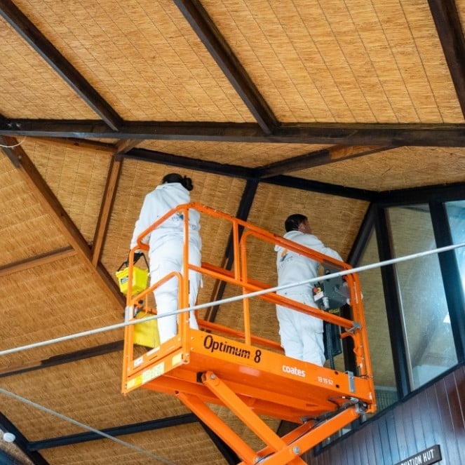Two workers in white uniforms stand on an orange scissor lift, meticulously conserving a ceiling