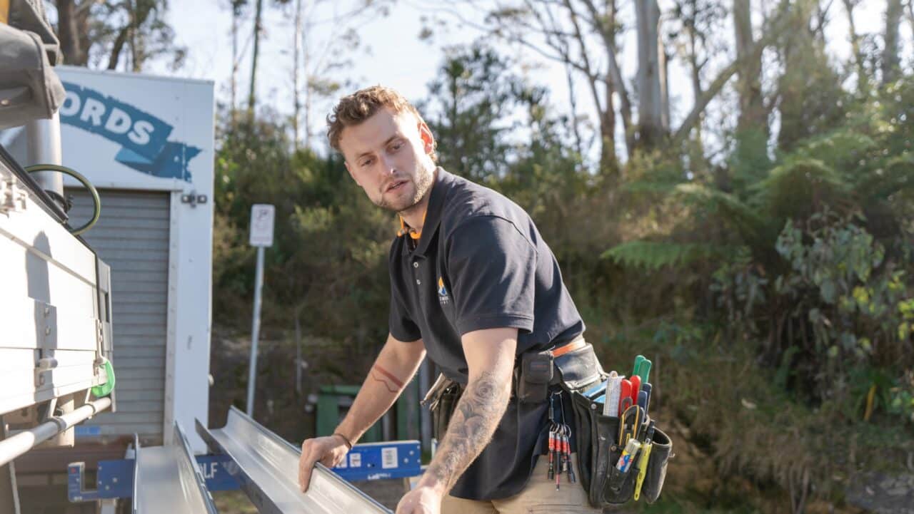 A man with a tool belt lifts a metal beam outdoors near machinery and foliage.