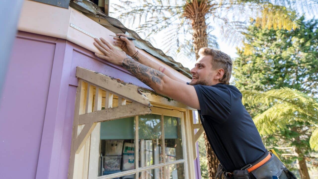 A man with tattoos measures a purple wooden structure with a ruler. Trees and sky are visible in the background.
