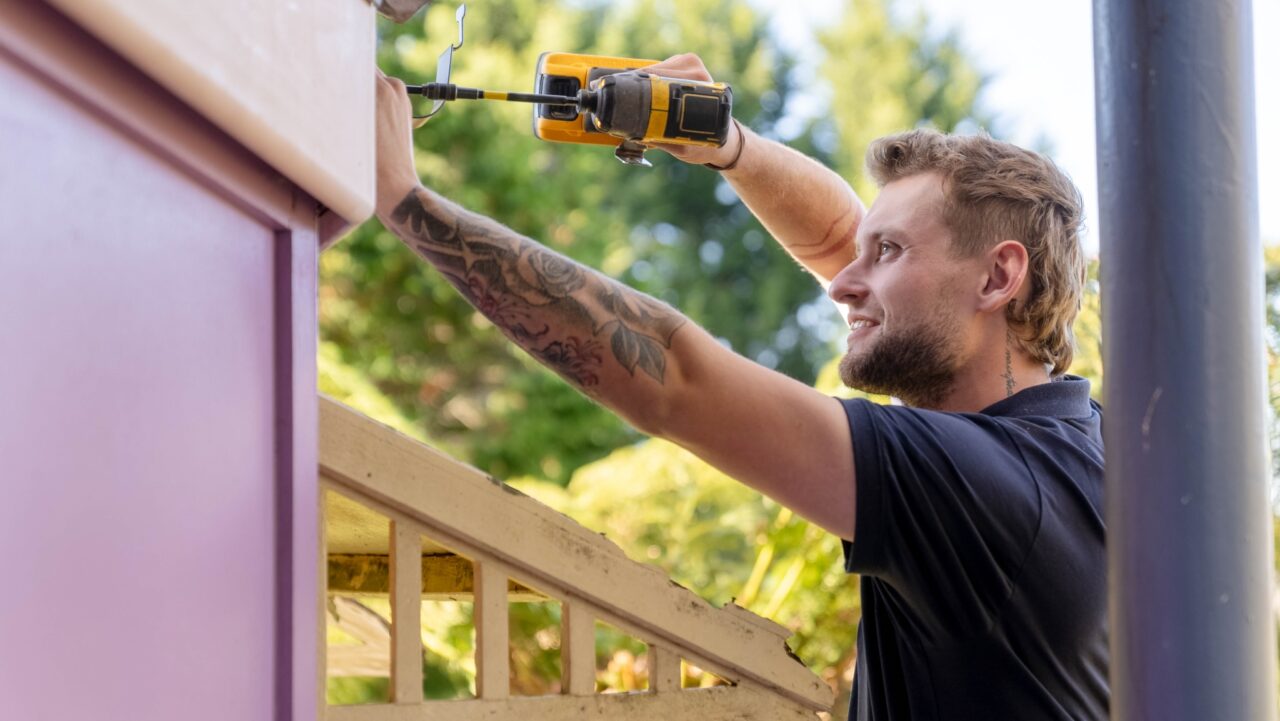 A man with tattoos on his arm, representing Expert Carpentry Services, uses a cordless drill to secure a structure outdoors in the picturesque Blue Mountains.