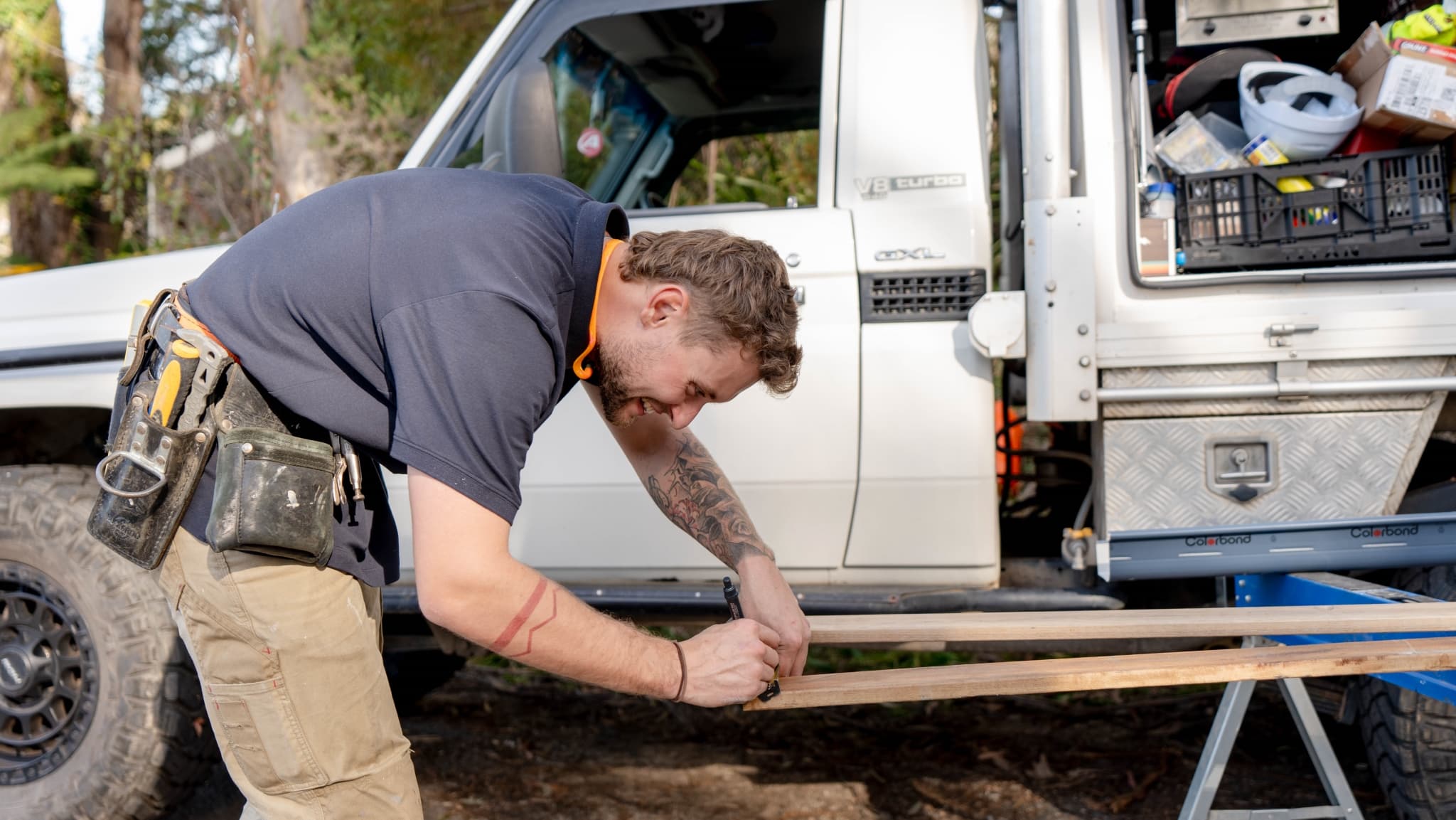 A man wearing a tool belt is measuring wooden planks outdoors next to a white utility vehicle with an open storage compartment.