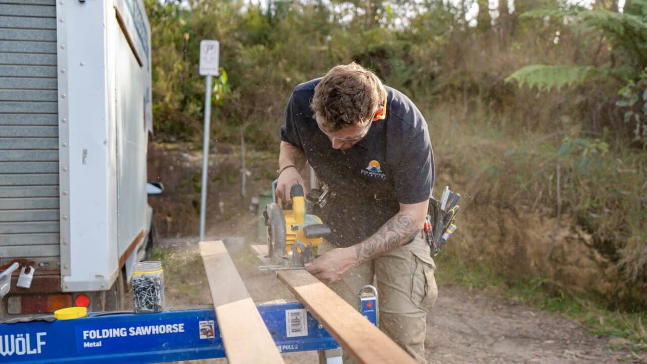 Person using a circular saw to cut wooden planks on folding sawhorses outdoors, with a toolbox in the background.