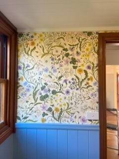 A wall with floral wallpaper featuring yellow, purple, and green patterns above blue wainscoting, next to a wooden window frame.