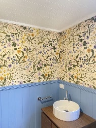 A small bathroom with floral wallpaper, blue wainscoting, a white round sink on a wooden vanity, and a wall-mounted faucet.