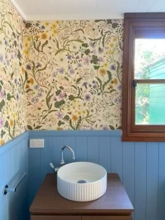 A bathroom with floral wallpaper and a blue wainscot panel. The sink has a round white basin and a silver faucet, positioned under a wooden-framed window with a green view outside.