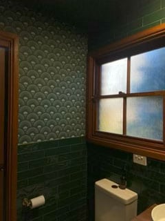 Bathroom with green fish scale pattern wallpaper, green tile wainscoting, a frosted window, toilet, and toilet paper holder on the wall.