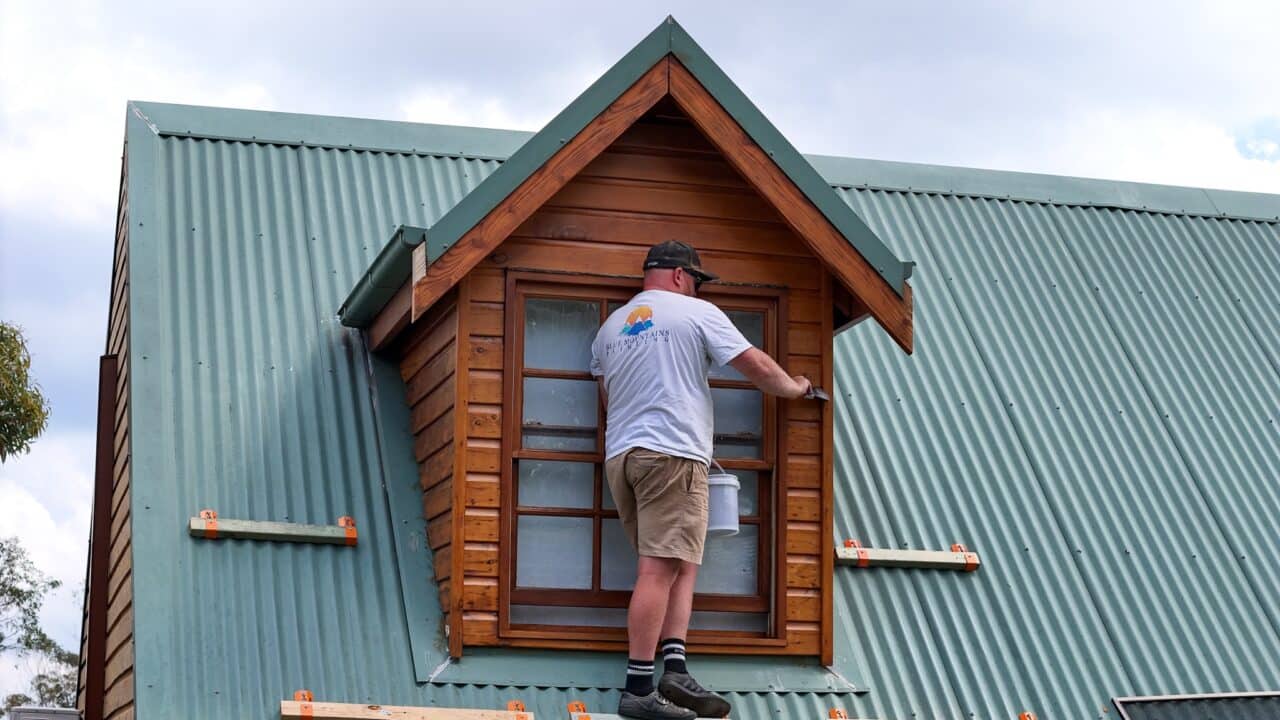 Person wearing a cap and white shirt painting a wooden dormer window