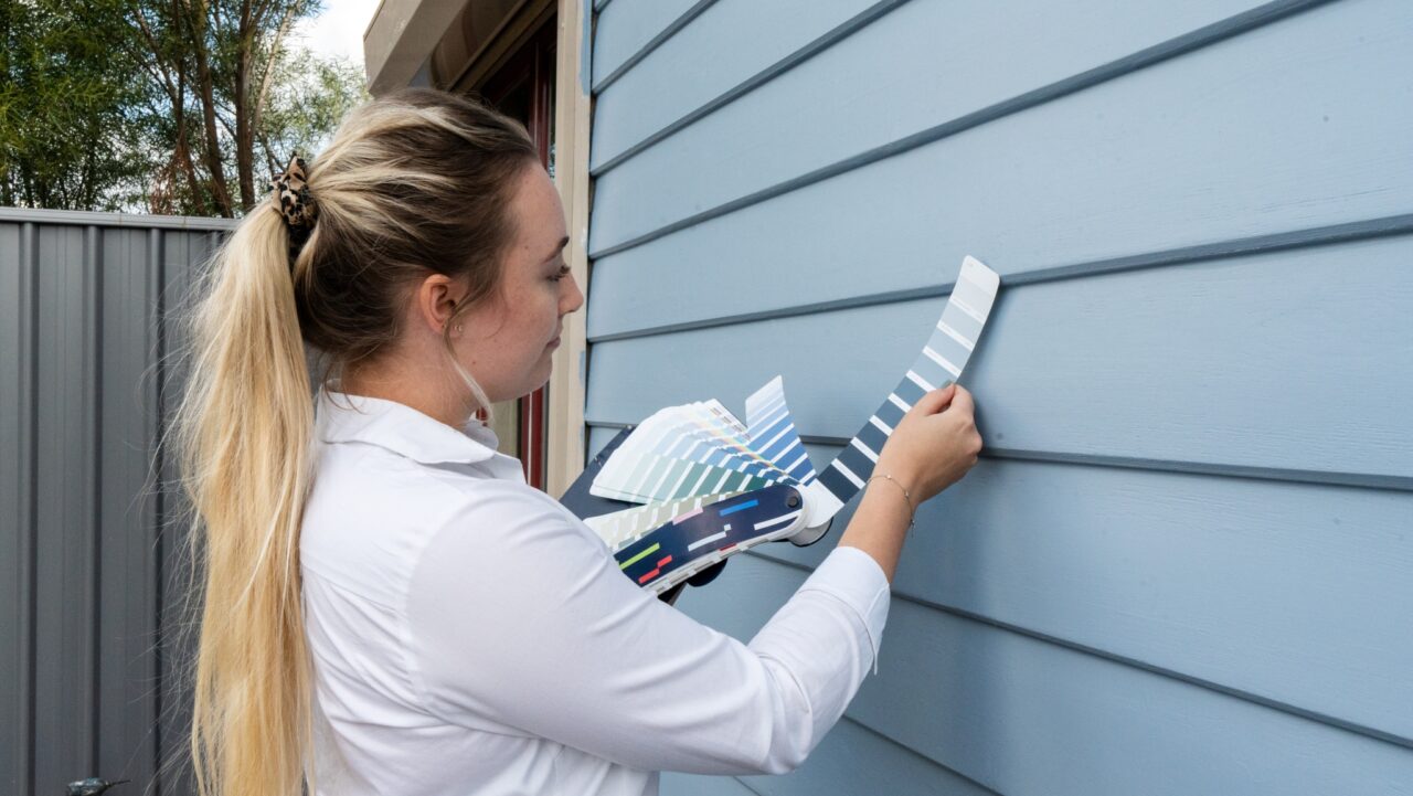 Person holding paint swatches against a blue house exterior to match colors.