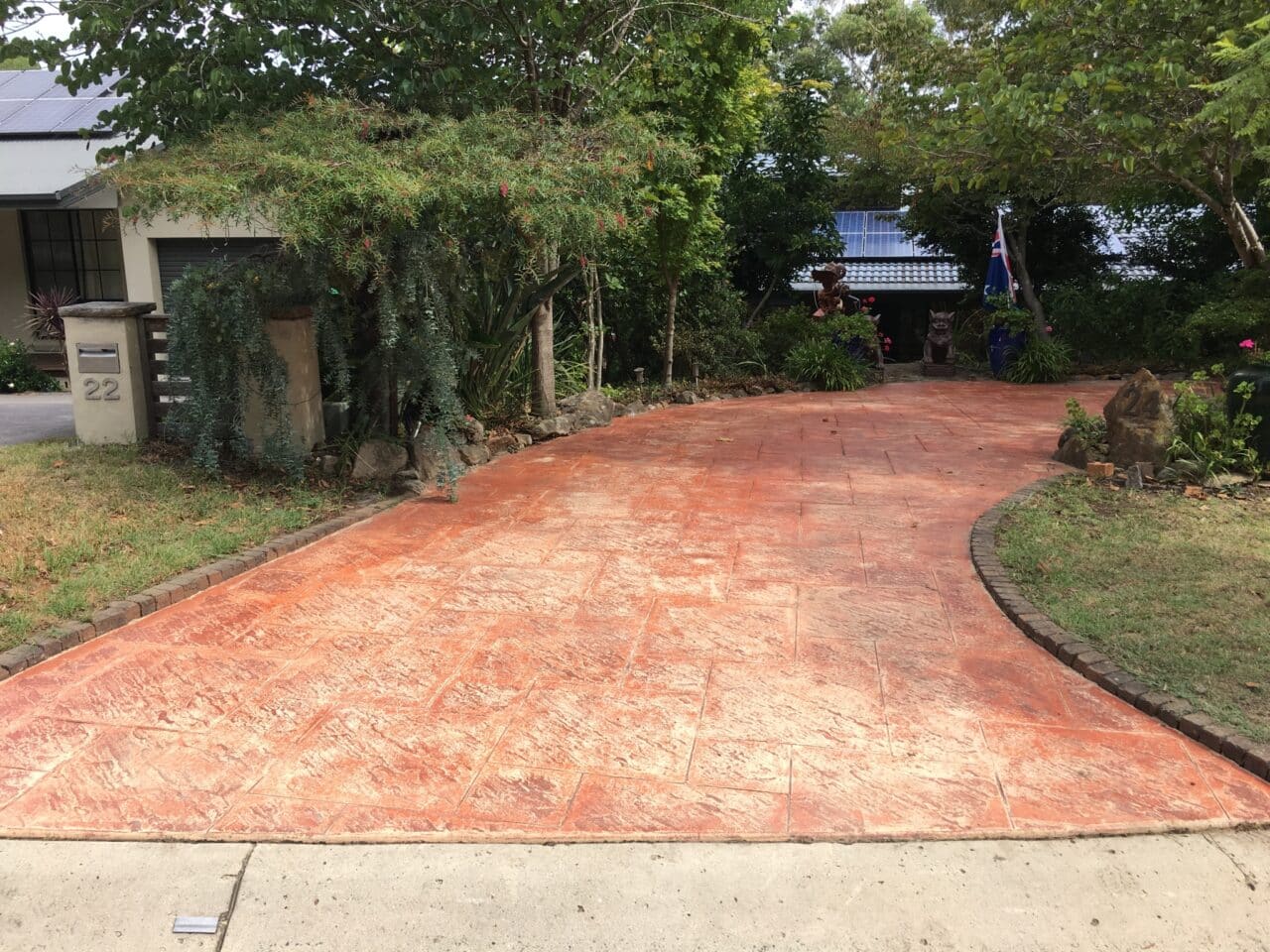 A curved stamped concrete driveway with a terra cotta color leads to a house surrounded by trees and greenery.