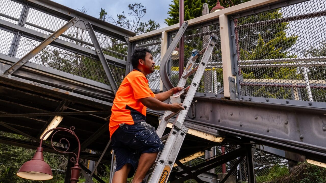 A worker in an orange shirt ascending a ladder to paint the metal framework of the Mt Vic bridge