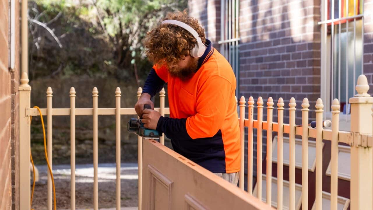 Person wearing an orange and navy shirt uses a power tool on a wooden board outside