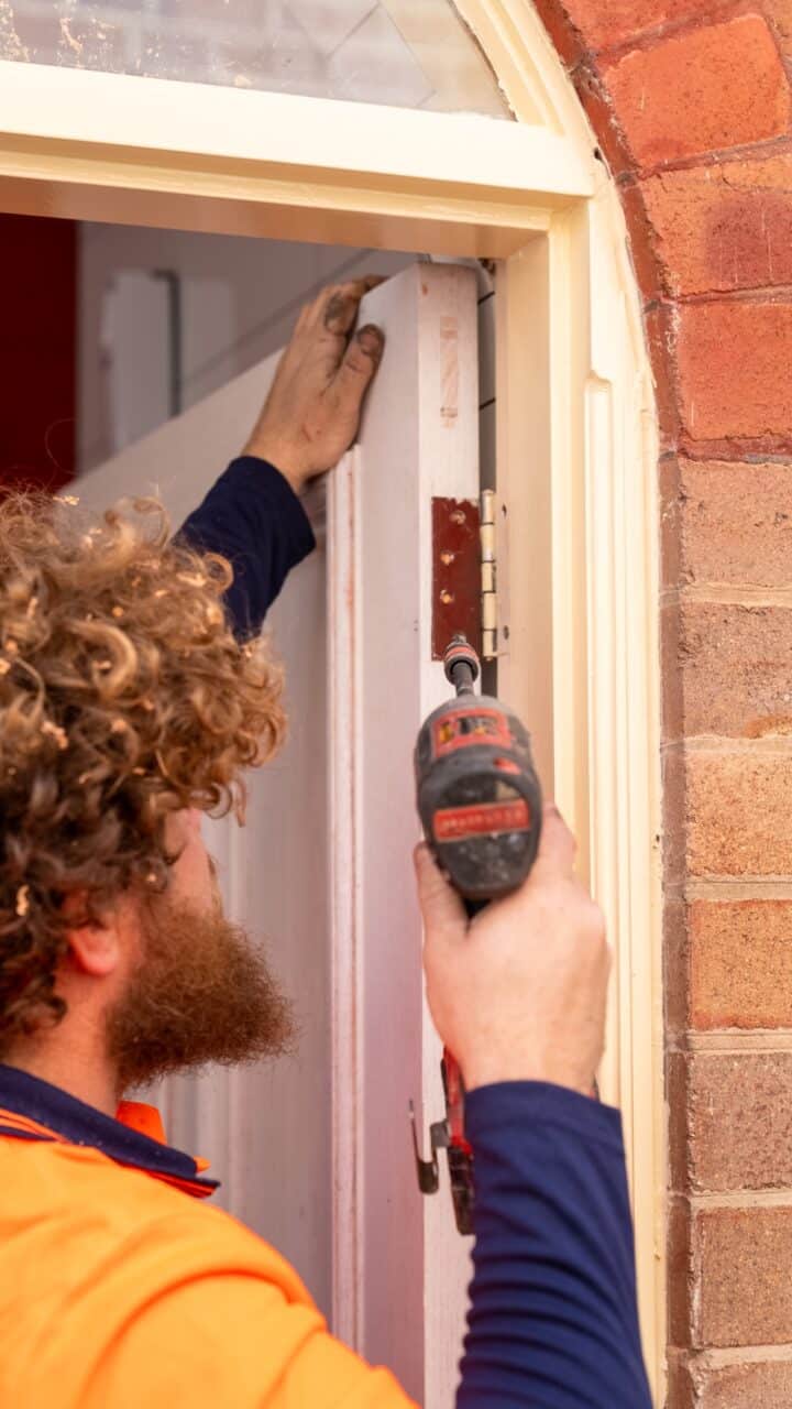A worker with curly hair installing a door hinge using a power drill near a brick wall