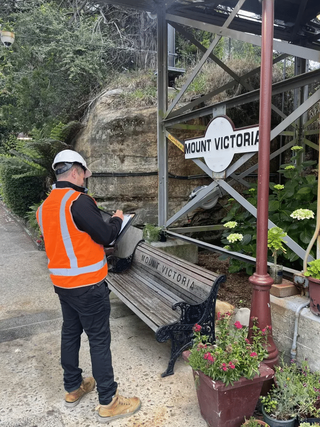 Person wearing an orange safety vest and white helmet writing on a clipboard near a bench