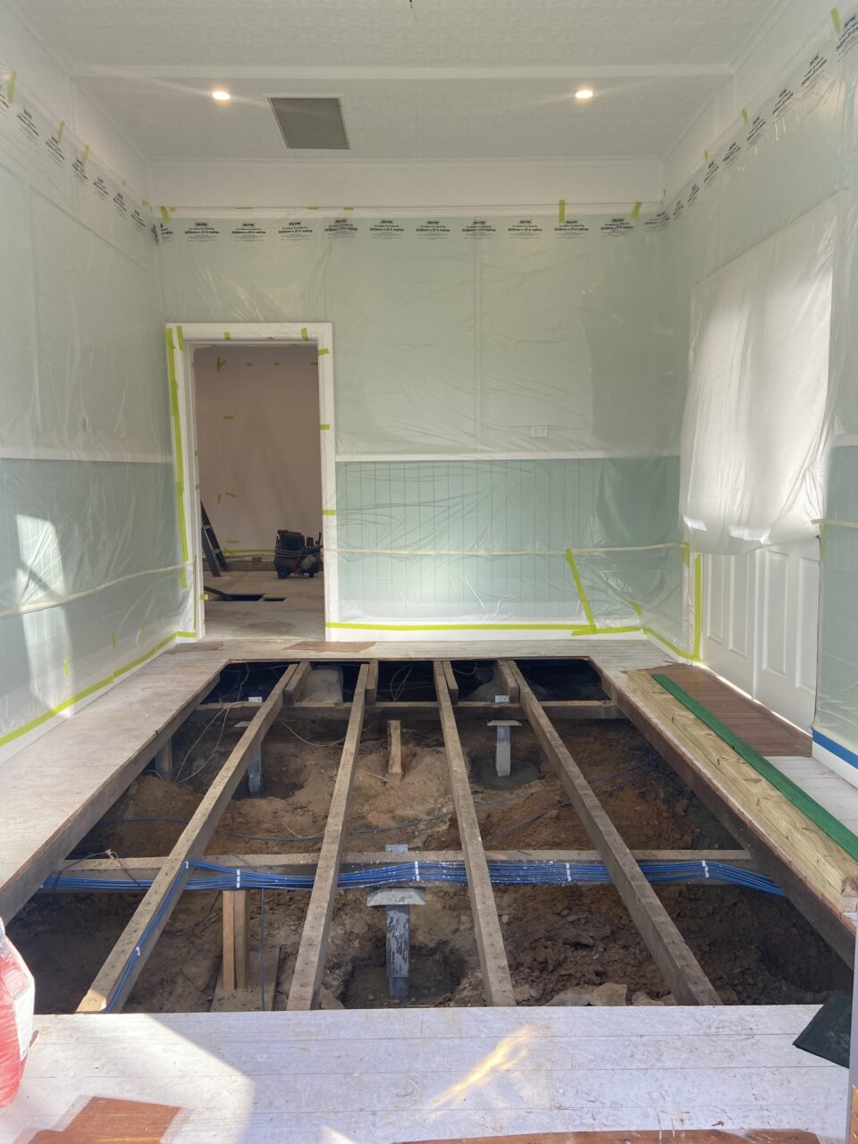 A room under renovation with walls covered in plastic sheeting and the floor removed, revealing exposed wooden beams and soil. A doorway leads to another room in the background.