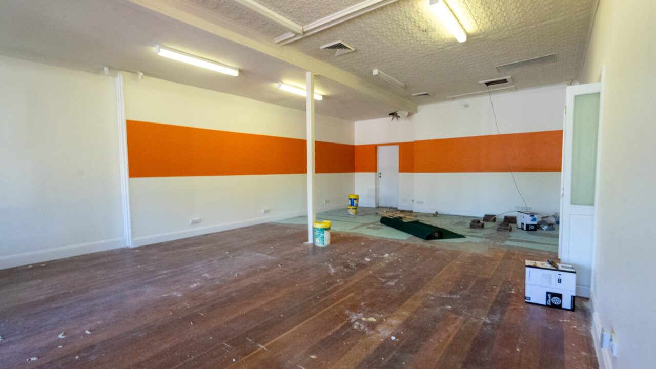 An empty room with wooden floors, white walls, and an orange horizontal stripe. Ceiling lights are on, and paint supplies are on the floor.