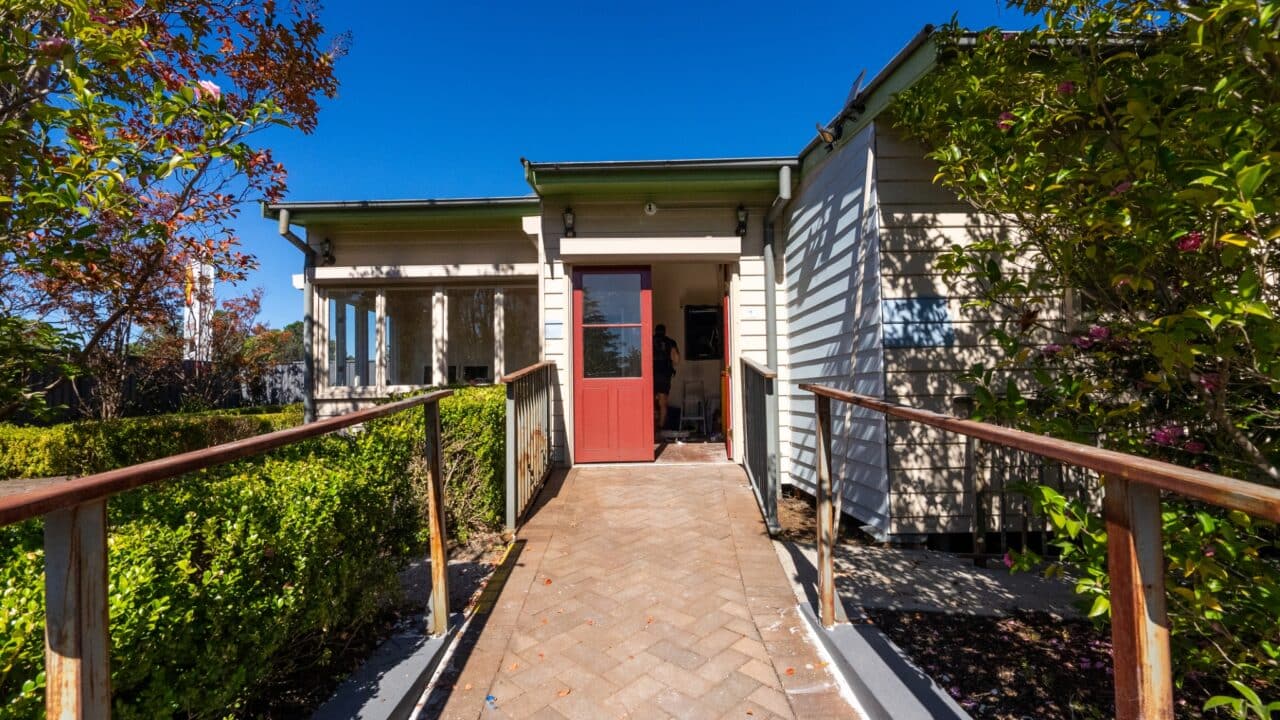 Wooden ramp leading to a house with a red door, surrounded by greenery under a clear blue sky.