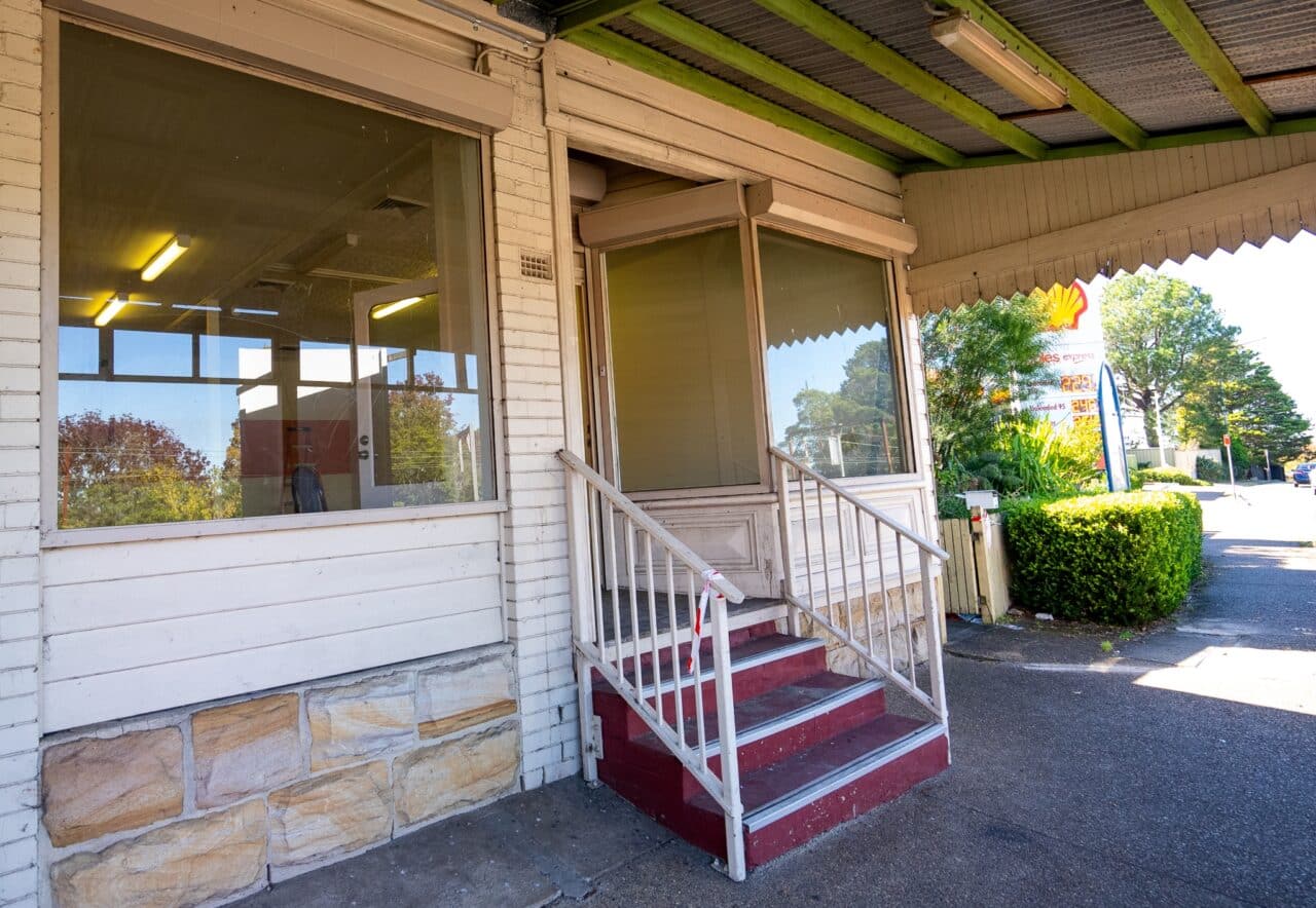 A small, closed shop with large windows and a red-painted step, located on a sidewalk with greenery visible in the background.