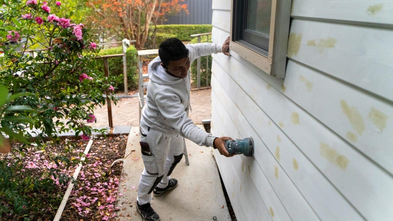 Person in white overalls sanding a house's exterior wall near a garden.