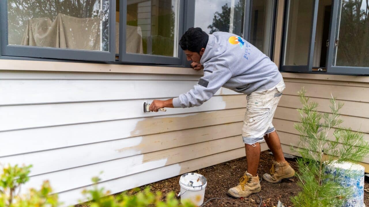 A person is painting the exterior of a house white, wearing a gray hoodie and beige shorts. A paint can is on the ground nearby, with a brush in use. Bushes are visible in the foreground.