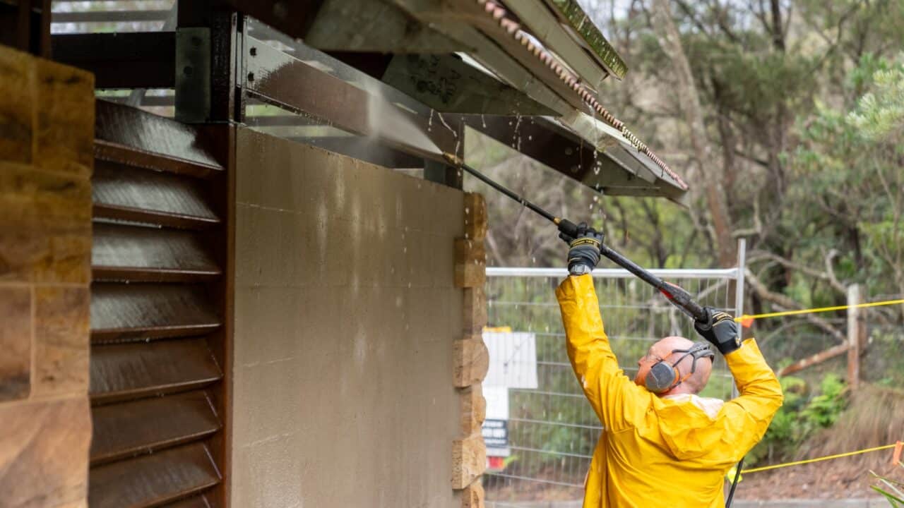 Person in a yellow raincoat and protective gear using a power washer to clean the exterior wall