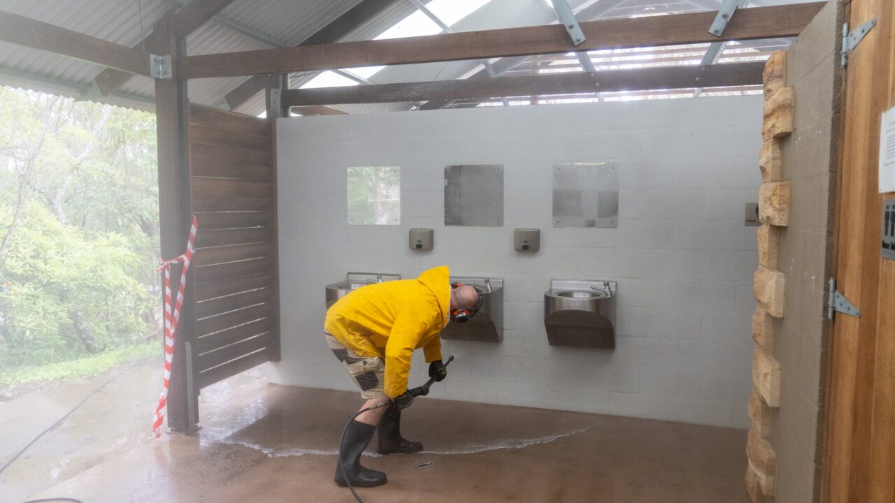 Person in a yellow raincoat and boots cleans a public restroom wall with a hose under a metal roof.