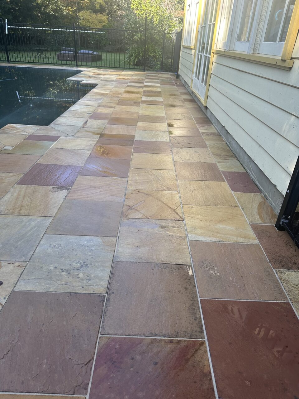 A patio with a multicolored tile pathway alongside a pool and a building with glass doors.
