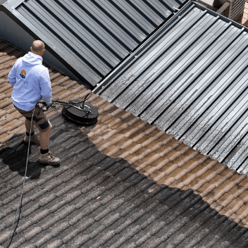 A person wearing a hoodie cleaning the corrugated metal roof of a hut with circular pressure washer