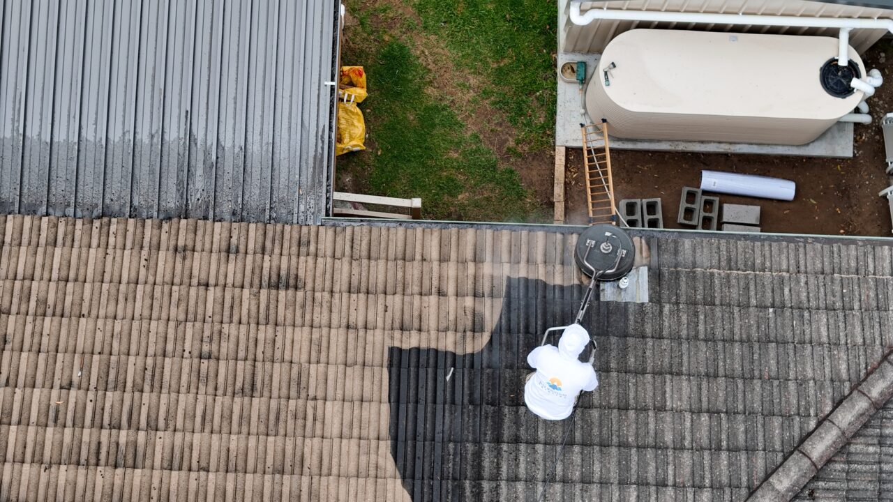 A person in protective clothing pressure washing a tiled roof, creating a visible clean area