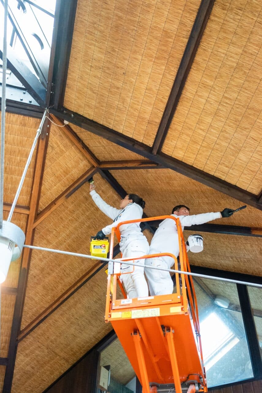 Two individuals in white overalls on an orange scissor lift painting a wooden ceiling