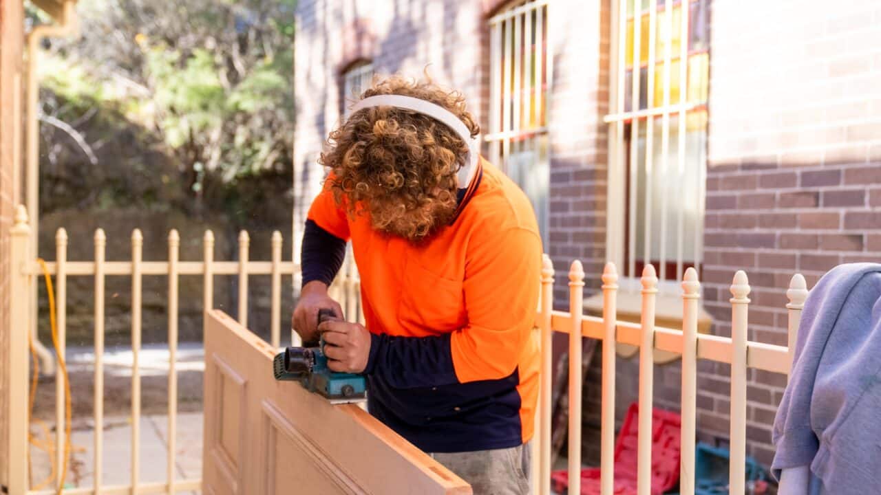 Person wearing orange safety shirt and earmuffs using a power tool to sand a wooden door outdoors.