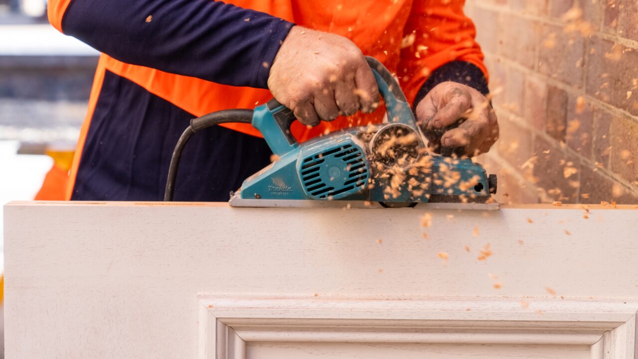 A person uses an electric planer to smooth the edge of a wooden door, creating wood shavings.
