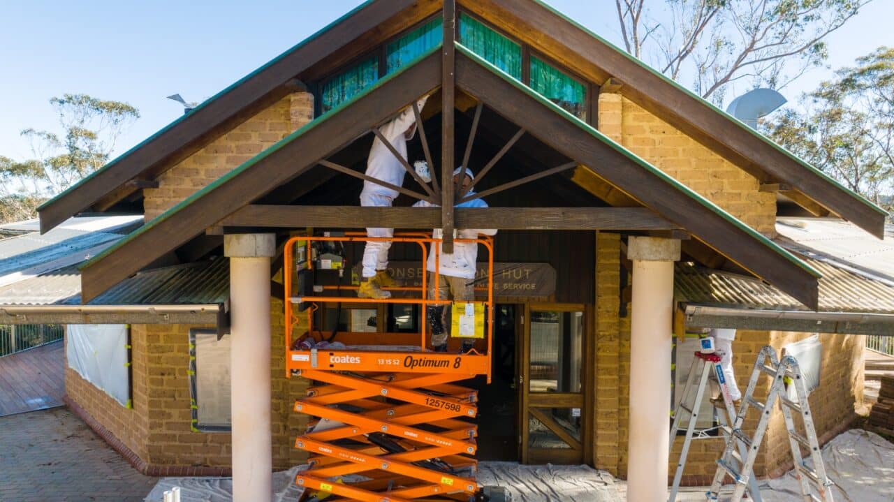 Two workers in white protective suits using scissors lift to conserve the exterior of a house.