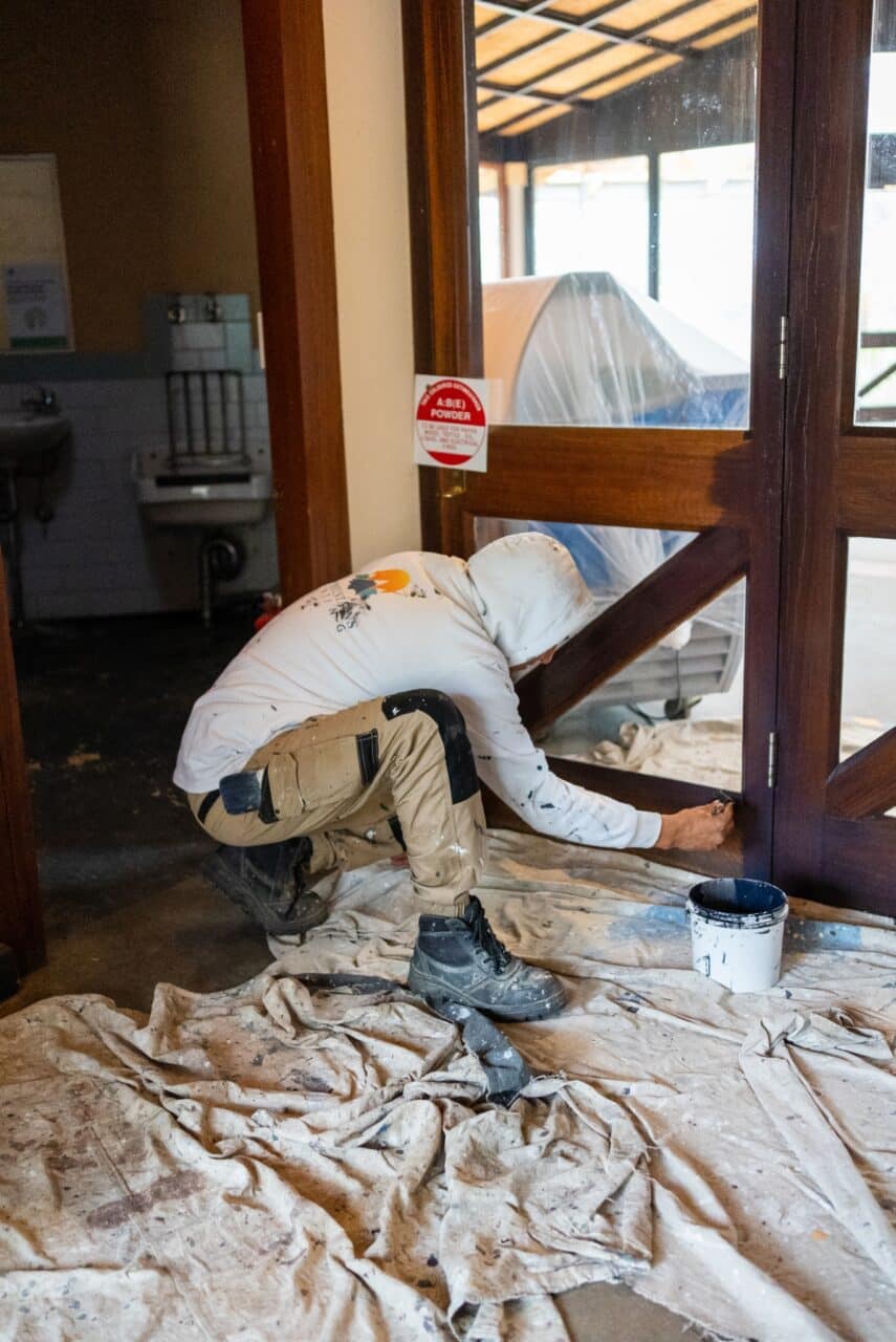 A person in work clothes crouches and painting a lower part of a wooden hut door.