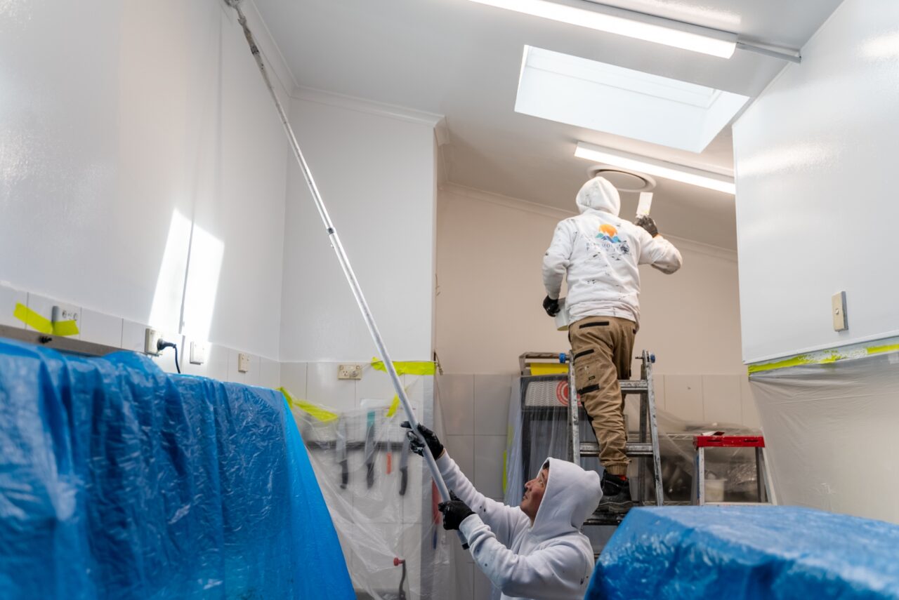 Two painters wearing white hoodies work on painting a high ceiling in a room.