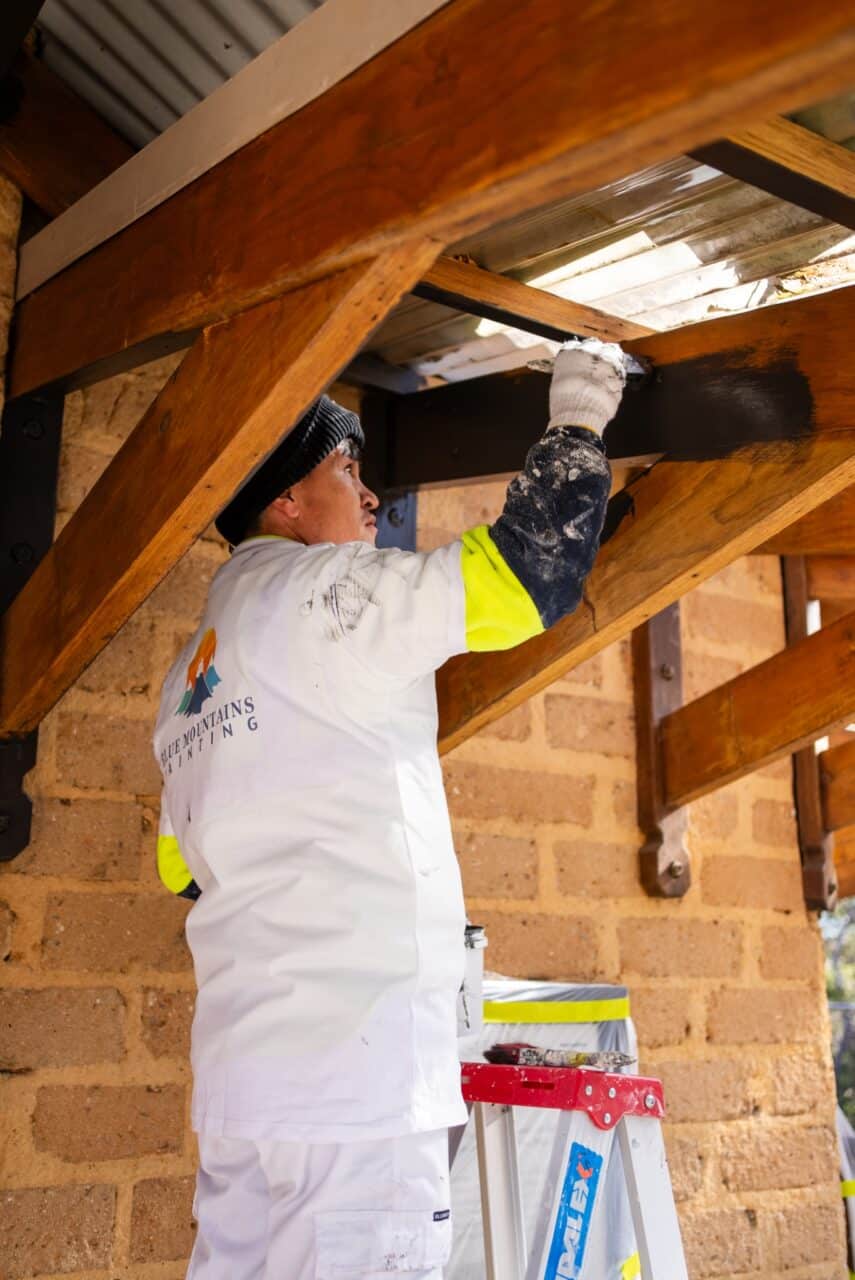 A worker in protective clothing stands on a ladder, painting a wooden beam.