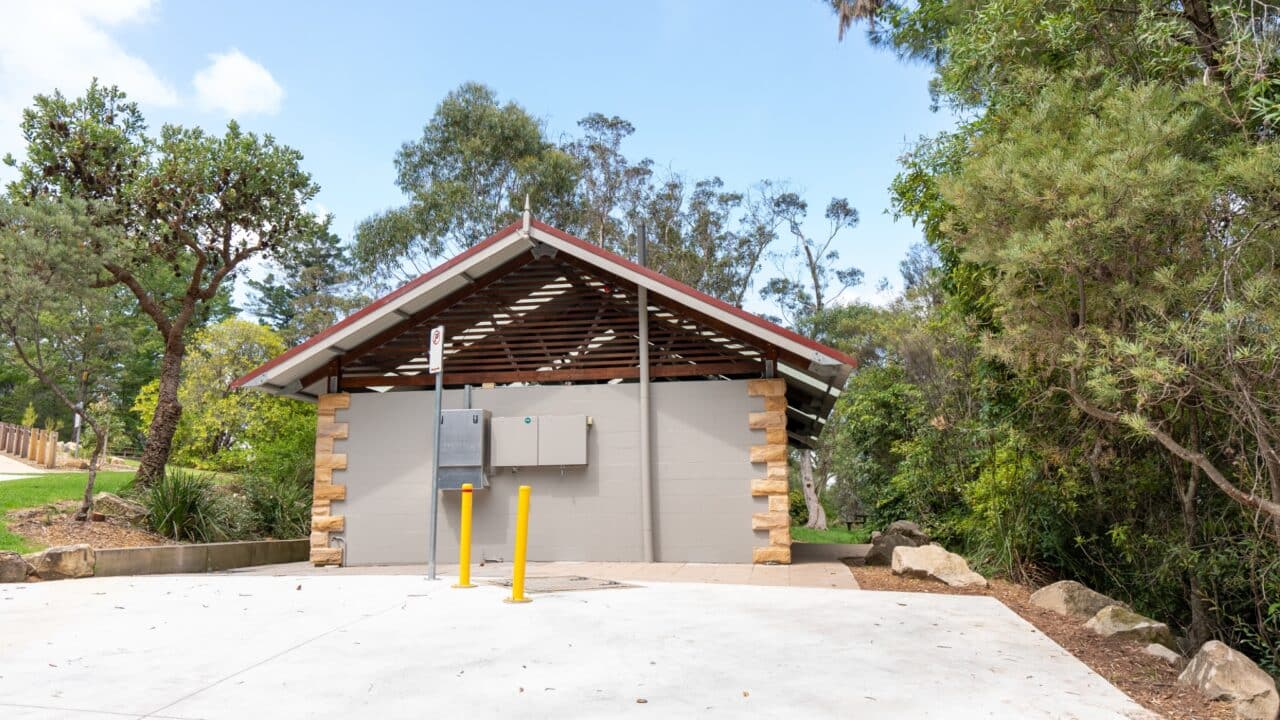 A small public restroom facility with a sloped roof, surrounded by trees and shrubs