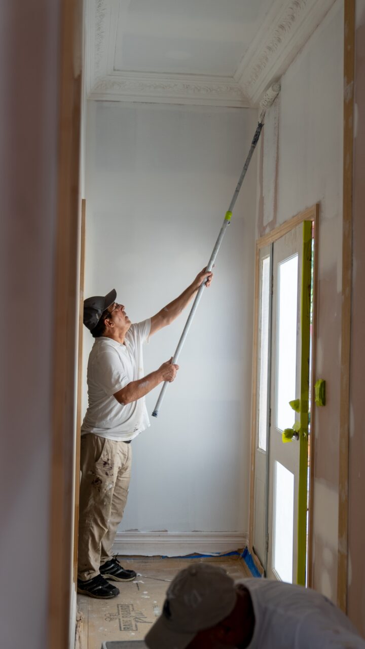 Man in a white shirt and cap using a long-handled roller to paint the top of a wall in a hallway