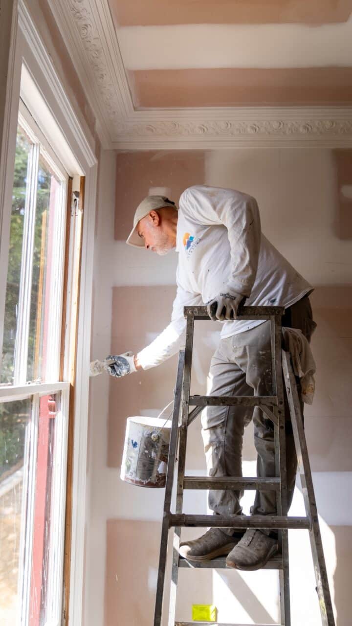 A person standing on a ladder and painting the trim of a window in a room