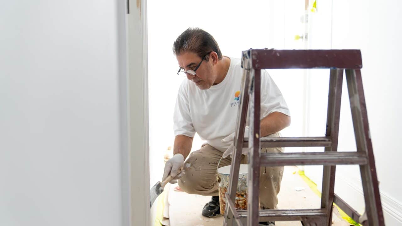 A man in a white shirt and gloves kneeling to paint a wall near a stepladder in a bright room