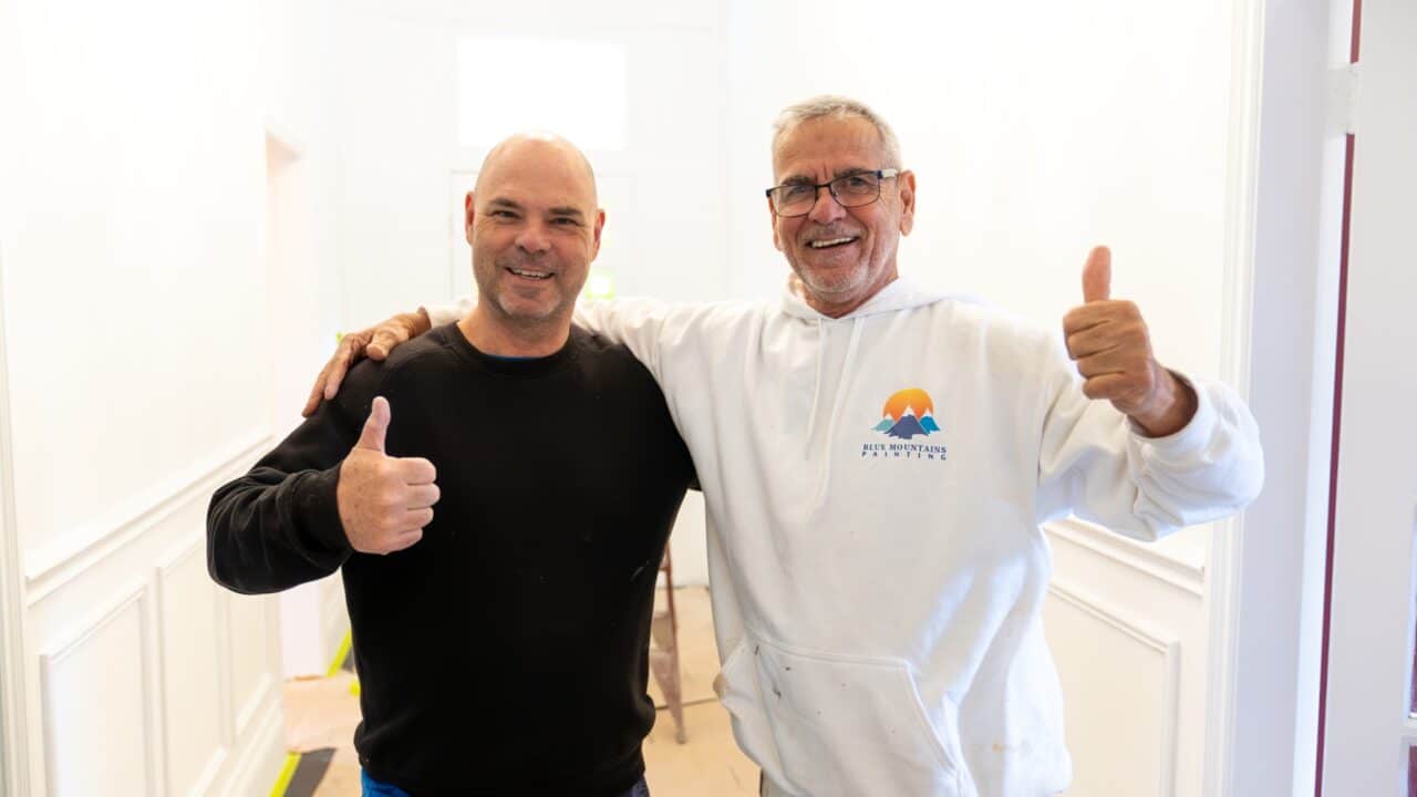 Two men standing side by side in a white-walled room, smiling and giving thumbs up