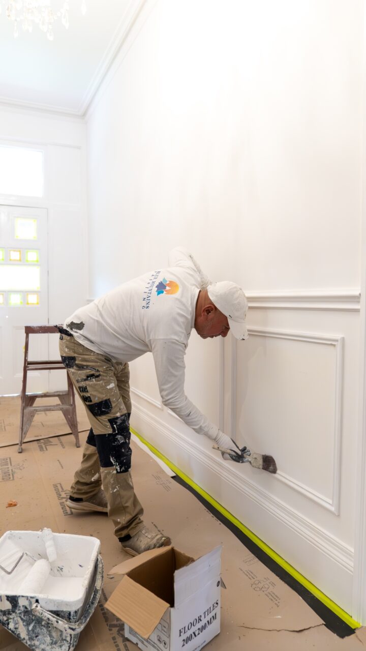 A painter wearing white work clothes applying paint to a white wainscoted wall