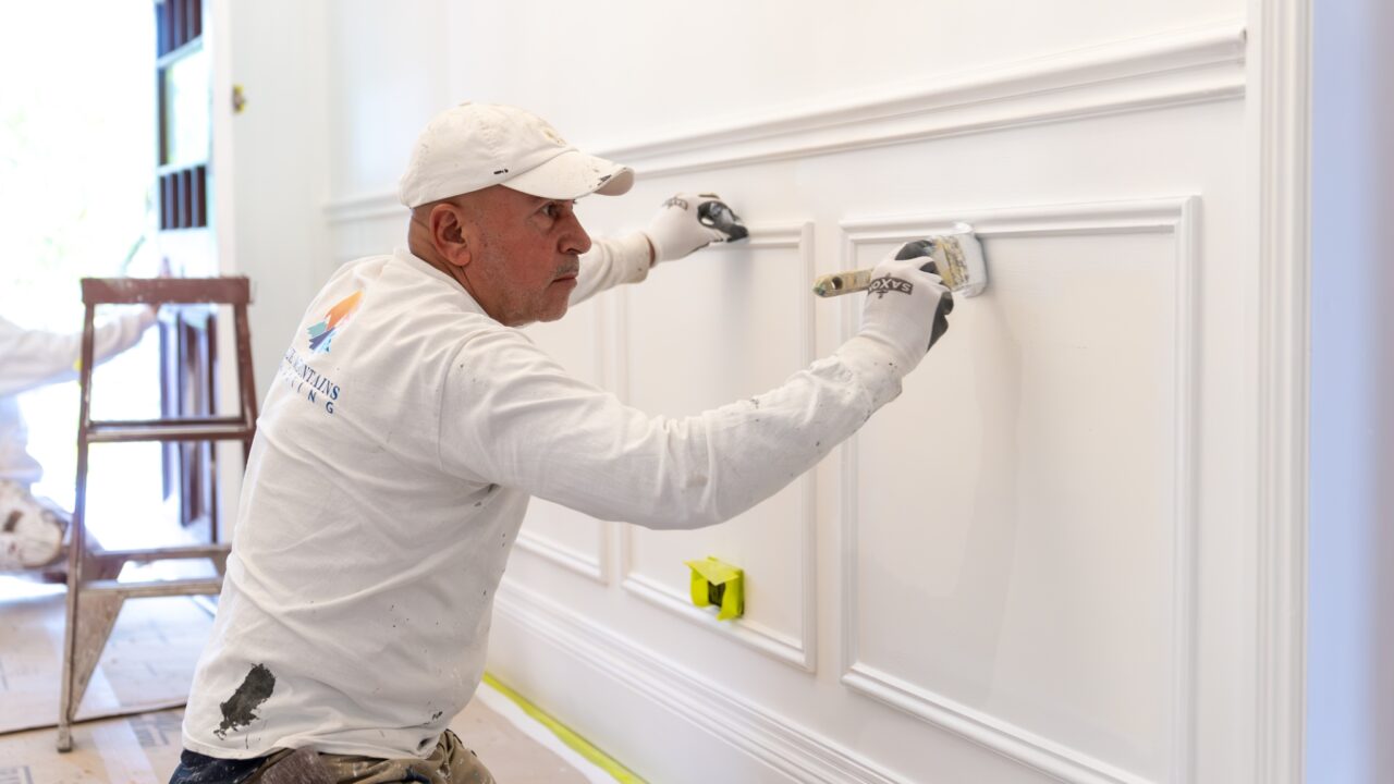 A man wearing a cap and gloves painting a decorative moulding on a white wall