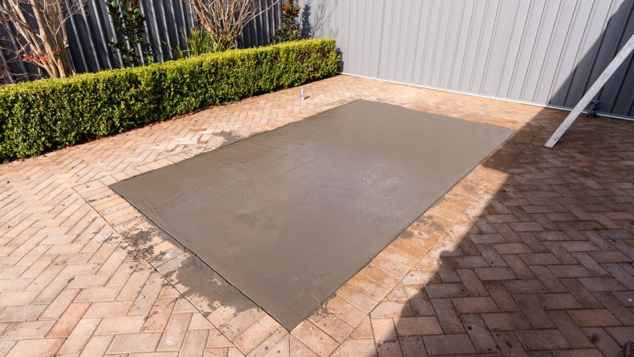 A large rectangular slab of freshly poured concrete on a herringbone-patterned brick patio, adjacent to a hedge and metal fence.