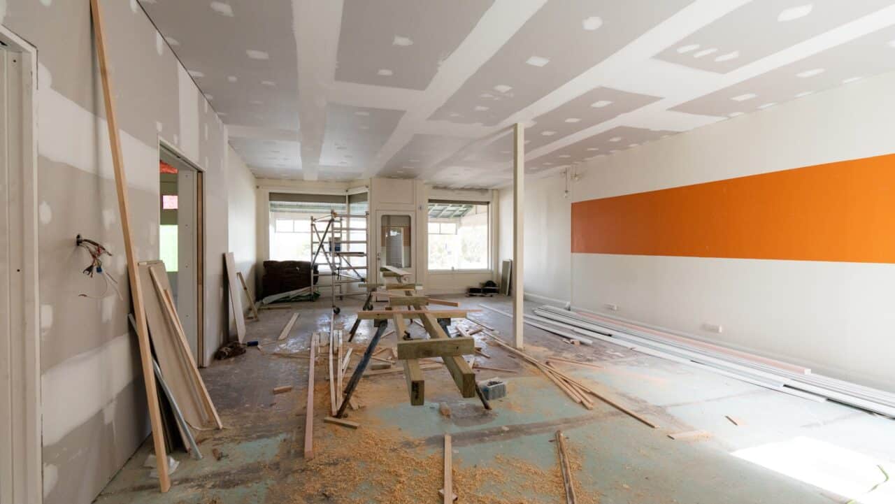 Interior of a room under renovation with construction materials, ladders, and tools scattered across the floor. Walls are partially painted with an orange stripe on one side.