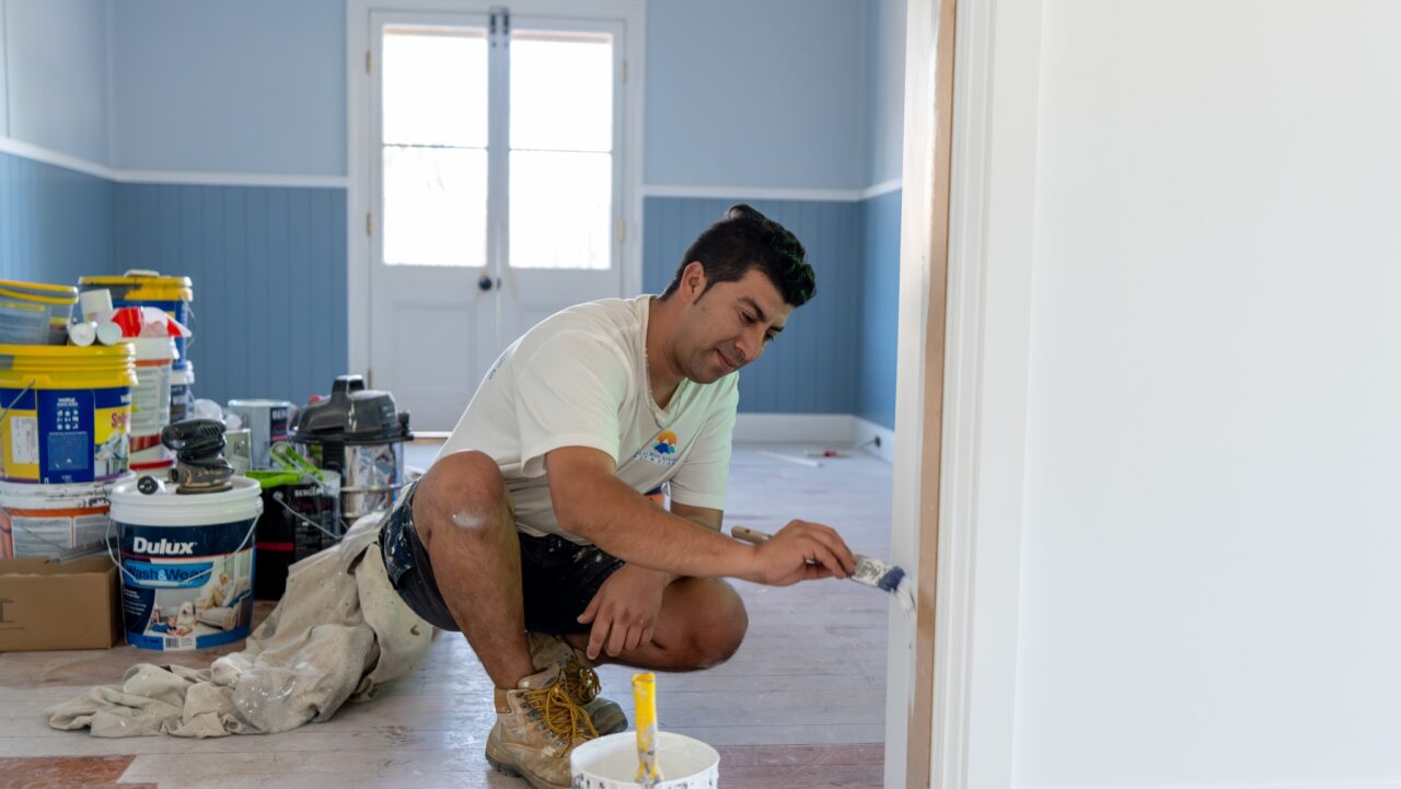 Person kneeling while painting a door frame in a room with blue walls and various painting supplies scattered around.
