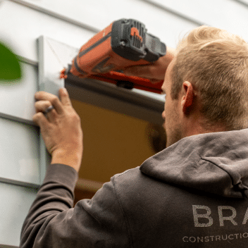 A person in a grey hoodie using a power nail gun to fasten siding near a window frame