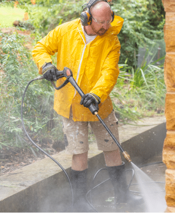 Person in a yellow raincoat, headphones, glove & shorts using a pressure washer to clean outside