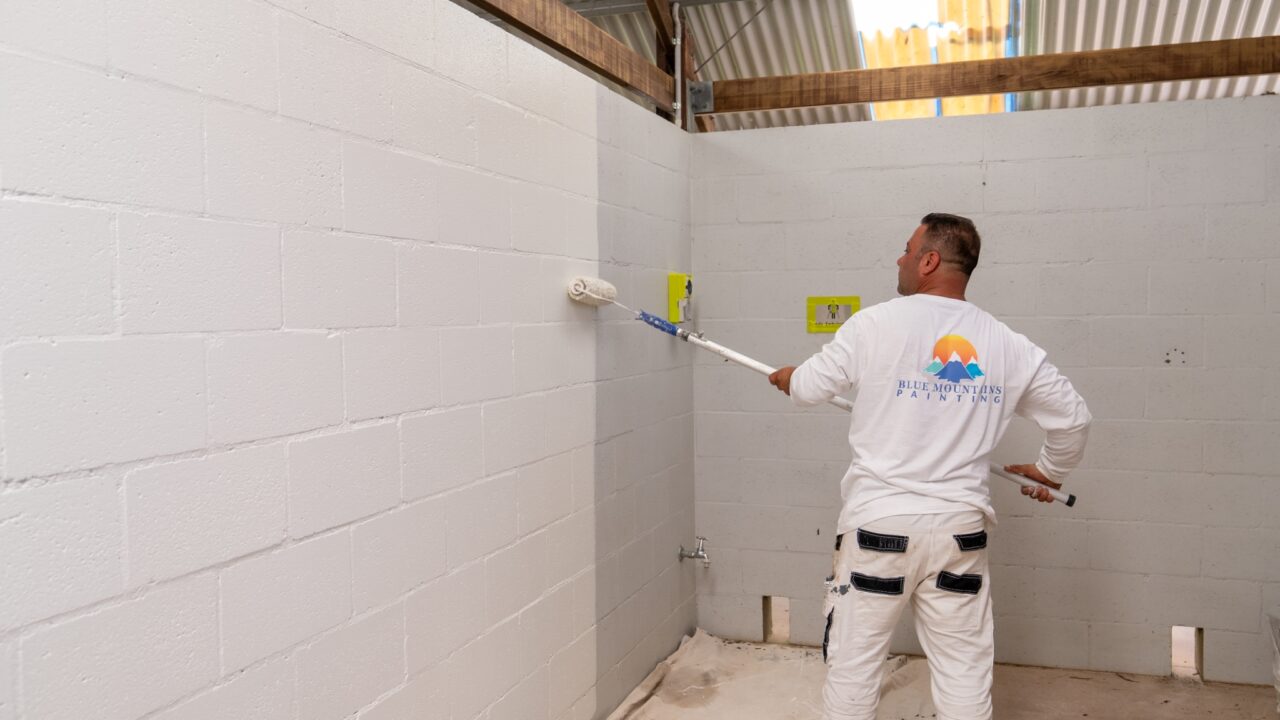 A man in a white shirt and pants painting a cinder block wall with a roller